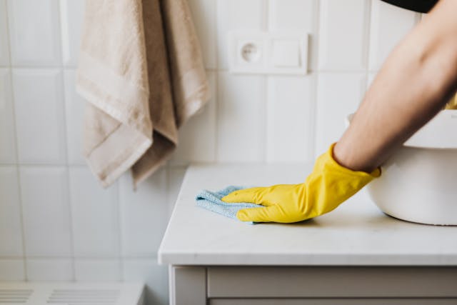A hand in a yellow rubber glove wipes down a bathroom surface